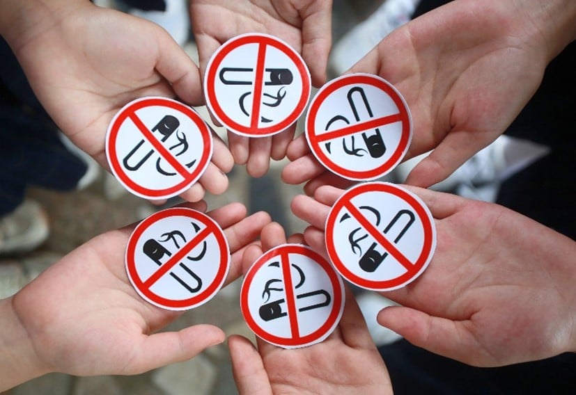 A group of hands holding red and white circular stickers with a prohibited symbol over a black and white drawing of a cigarette.